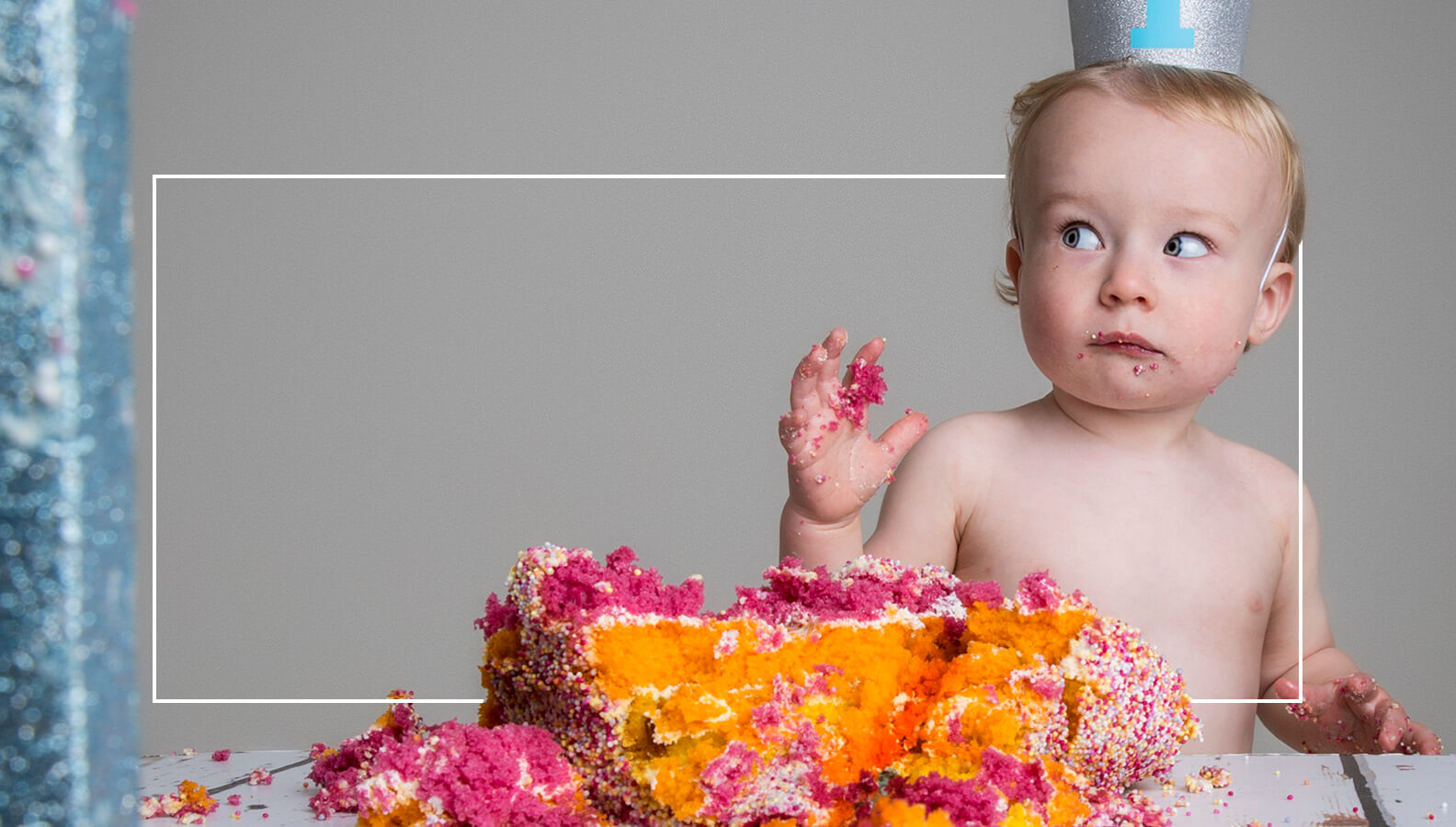 Image of one year old smashing a cake with crumbs all over his hands and checking to see if Mummy has seen